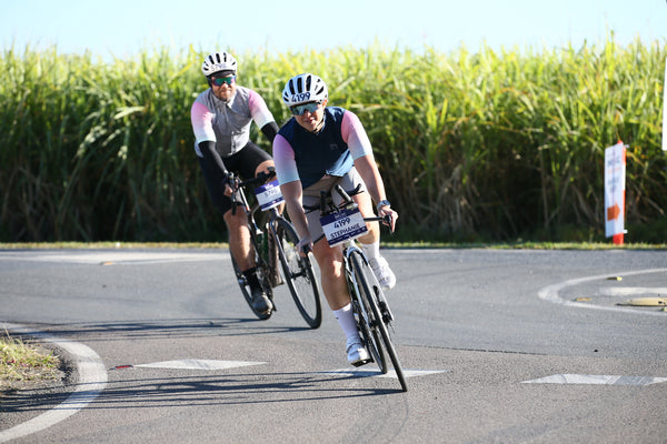 Two cyclists racing on a paved road with a tall green crop in the background. The cyclist in the foreground wears a black and white helmet, sunglasses, a pink and white jersey, and a bib number, while the second cyclist trails behind in a pink and white jersey.